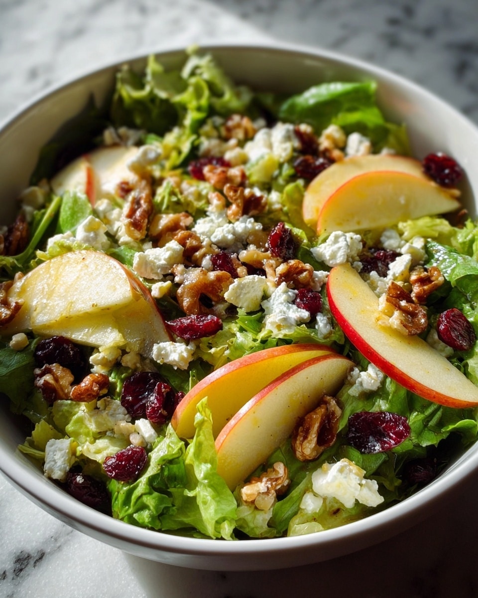 A white bowl filled with a colorful salad sitting on a white marbled surface, starting with a base layer of fresh green leafy lettuce. On top are thin light yellow apple slices with red edges, scattered evenly across the salad. White, crumbly cheese bits are spread all over, mixed with dark red dried cranberries and medium brown walnuts that add texture and warmth. The lighting highlights the fresh, natural colors of the ingredients, creating an inviting look. photo taken with an iphone --ar 4:5 --v 7