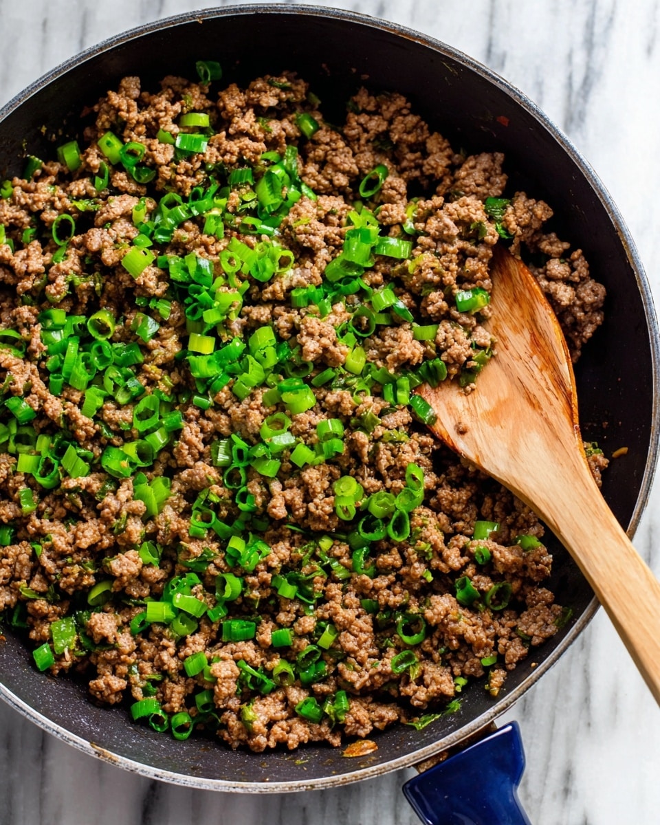 The image shows a pan filled with cooked ground meat mixed with bright green chopped scallions scattered evenly on top. The meat is crumbly and browned, covering the entire pan with some areas having slightly darker toasted bits. A wooden spoon is scooping a portion of the meat from the right side, its smooth texture and natural brown color contrasting with the meat. The pan has a dark interior and a small part of a blue handle is visible at the top and bottom edges. The background features a white marbled surface underneath the pan. photo taken with an iphone --ar 4:5 --v 7