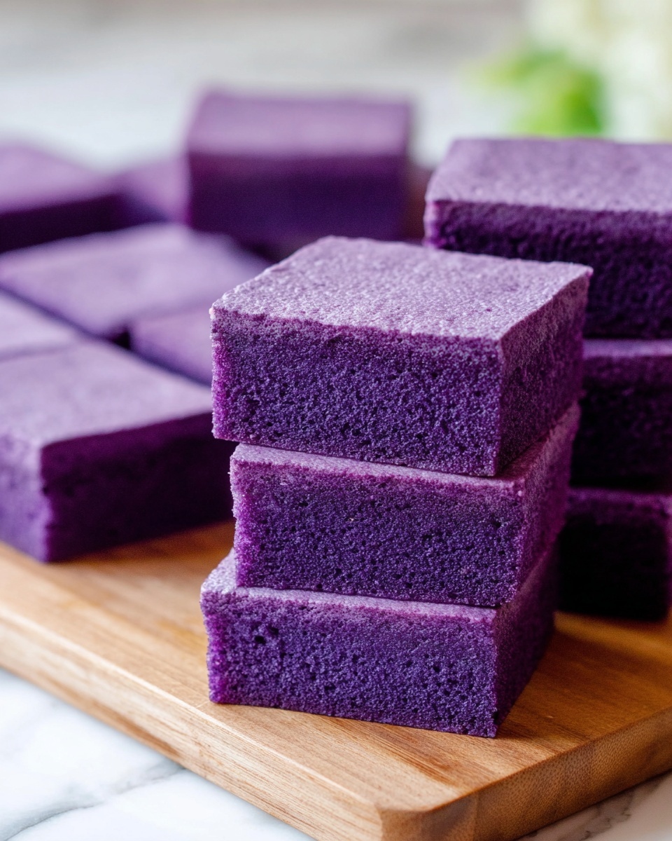 The image shows a group of purple square-shaped cakes neatly cut and stacked. The stack in front has three visible layers, each a solid purple color with a soft and moist texture. Behind the stack, there are more purple squares laid out flat in an organized way on a wooden surface. The purple color is rich and even throughout the cakes. The scene is set on a white marbled texture. Photo taken with an iphone --ar 4:5 --v 7