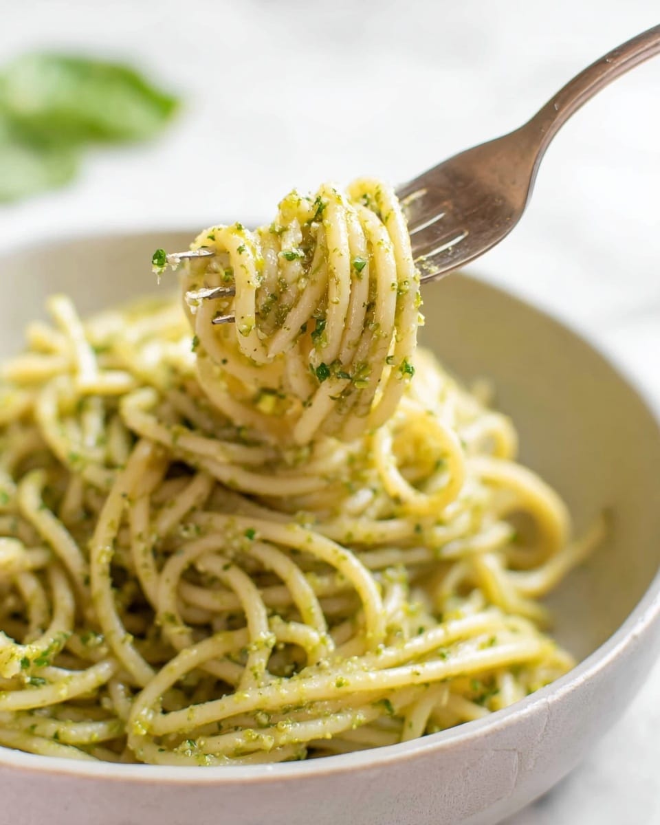A white bowl filled with twirled spaghetti noodles coated in a light green pesto sauce with small herb bits visible throughout. A silver fork lifts a small twirl of the spaghetti from the bowl, showing the noodles coated evenly in the sauce with a glossy texture. The bowl sits on a white marbled surface, with a faint green leaf blurred in the background. The photo is close-up and focused on the fork and pasta strands, showing the smooth and slightly oily texture of the sauce on the pasta. photo taken with an iphone --ar 4:5 --v 7