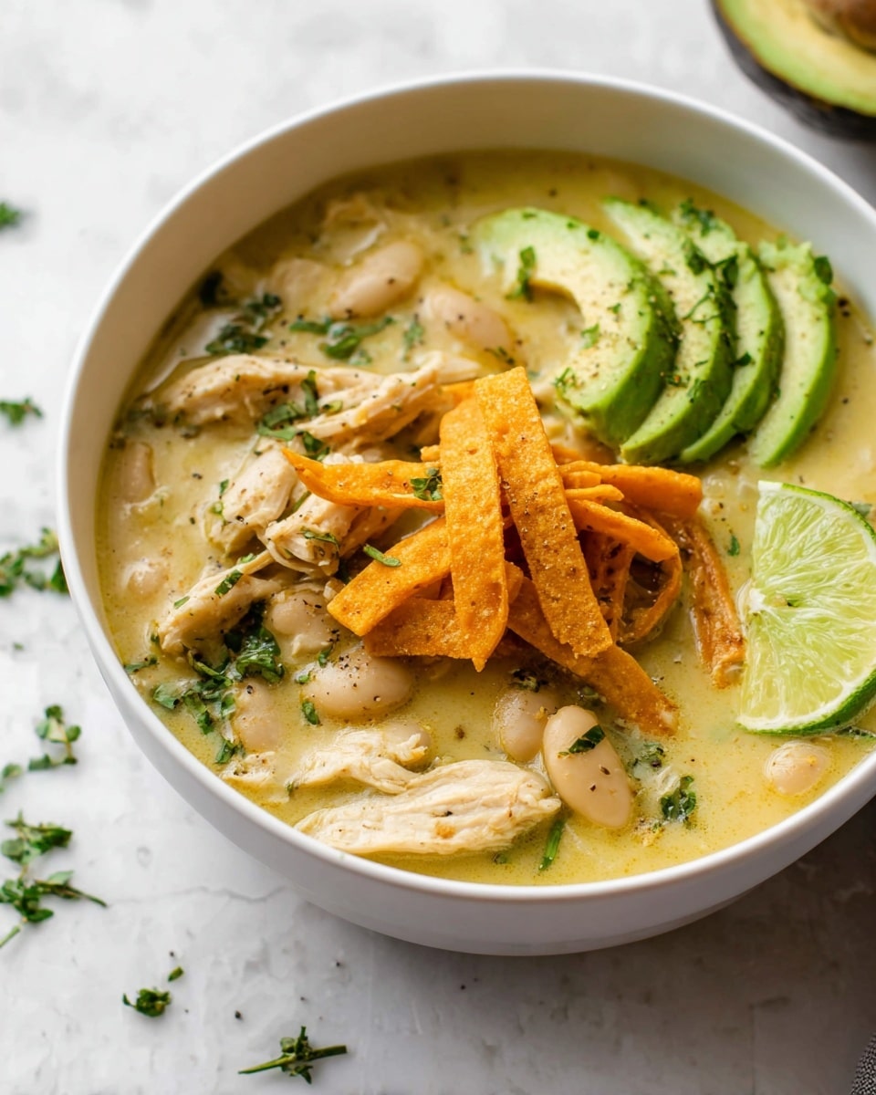 A white bowl filled with creamy yellow soup that has pieces of cooked white beans and soft light brown chicken slices in it. On top of the soup sits a small pile of crispy, bright orange fried tortilla strips. To the side of the bowl there are three fresh light green avocado slices sprinkled with black pepper and some chopped green herbs scattered on the soup and around the avocado. A wedge of lime with a pale green peel and juicy light green inside rests on the edge of the soup near the chicken. The bowl is placed on a white marbled surface, and a few small green herb leaves are scattered nearby. Photo taken with an iphone --ar 4:5 --v 7
