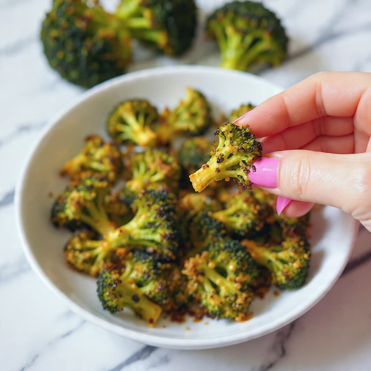 A white plate holds a pile of crispy broccoli pieces that are coated with a light brown crunchy layer, with the green broccoli florets peeking through. The broccoli looks cooked and slightly charred on top, giving a mix of dark and bright green colors. A woman's hand with pink nail polish is lifting one piece of broccoli with the crunchy coating. In the background, there are blurred green broccoli heads on a white marbled surface. Photo taken with an iphone --ar 4:5 --v 7