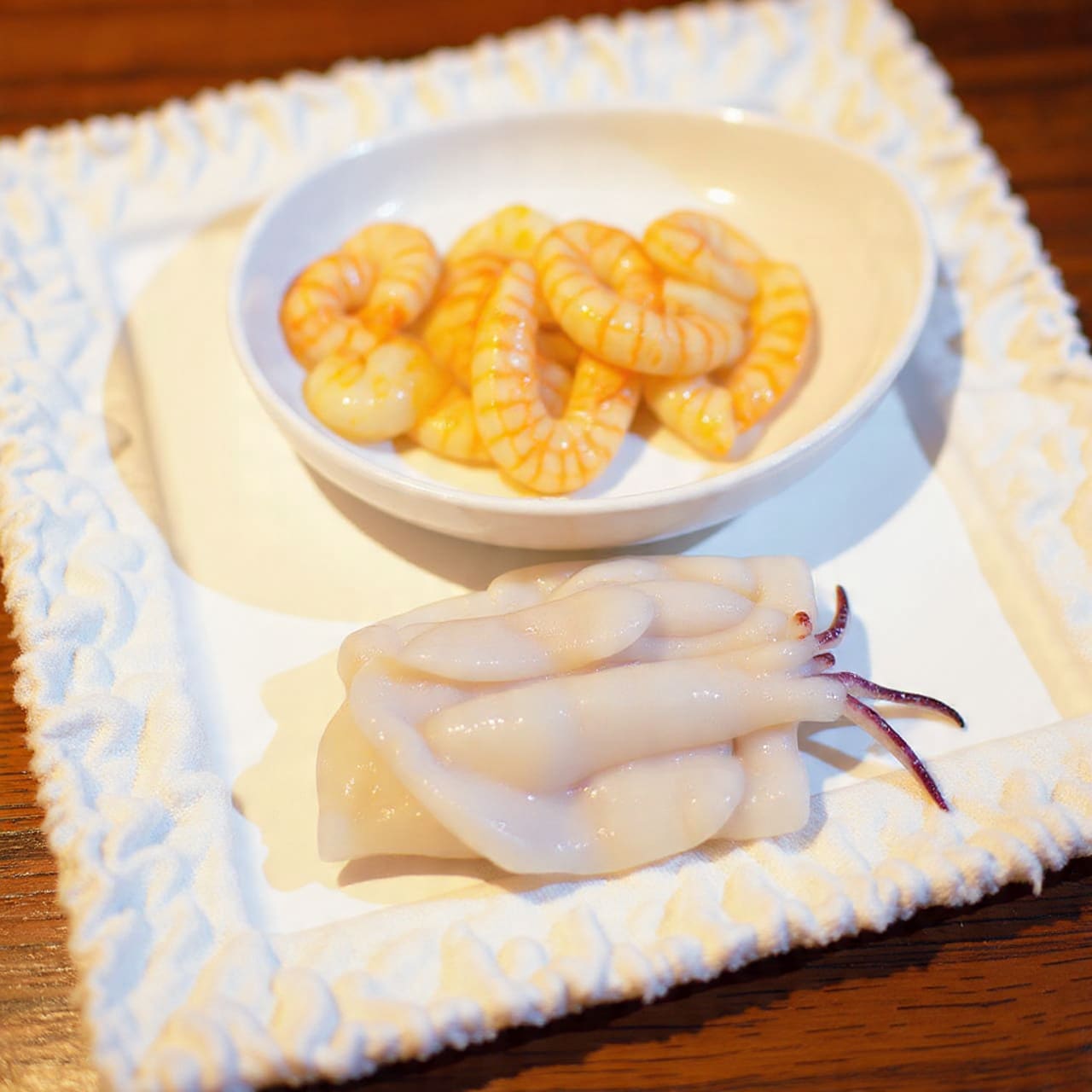 The image shows two layers of seafood laid out on a white textured cloth. The top layer is a group of small, light pink shrimp with a slightly shiny surface, covering about half of the area. The bottom layer consists of folded white squid pieces mixed with dark purple tentacles, creating a contrast with the shrimp above. Both layers are placed flat side by side on the cloth, which is set on a wooden surface with a warm tone. photo taken with an iphone --ar 4:5 --v 7