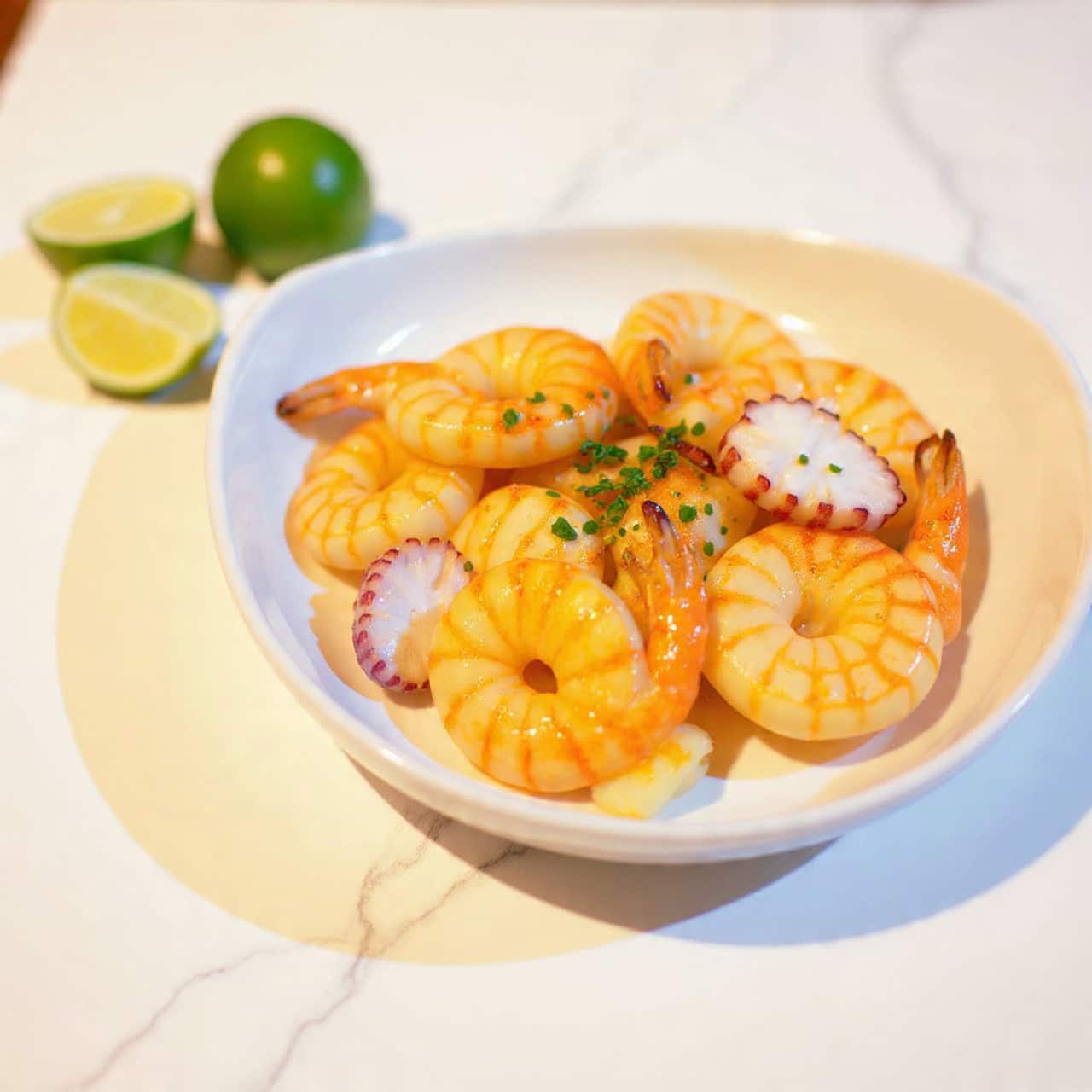 The dish is a white bowl filled with cooked shrimp and small octopus pieces, both showing a golden brown grilled texture with some bits of herbs sprinkled on top. The shrimp are plump, slightly curved, and mixed with small tentacle pieces of the octopus that have a purplish hue. There are two fresh lime wedges placed on the left side of the bowl adding a touch of bright green color. Small green herb leaves are scattered over the seafood, giving a fresh look. The bowl is placed on a white marbled surface. Photo taken with an iphone --ar 4:5 --v 7