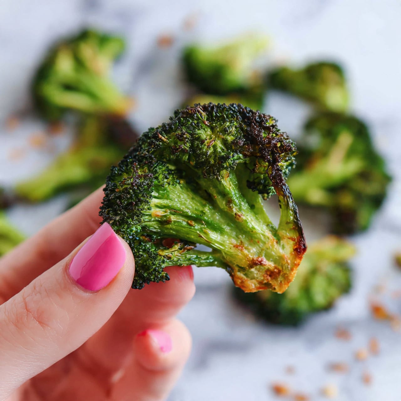 A close-up of a crispy broccoli chip held by a woman's hand with pink polish on the fingernail, showing the top layer of dark green roasted broccoli florets and a thin golden-brown crunchy base beneath with a few small holes. In the background, more broccoli chips are blurred on a white marbled surface scattered with crumbs. photo taken with an iphone --ar 4:5 --v 7
