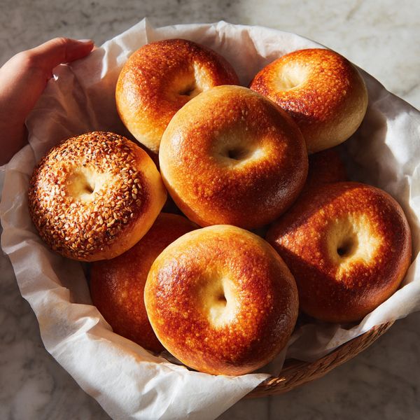 The image shows a basket filled with round bagels that have a shiny, golden-brown crust. Some bagels have a nut topping, adding a slightly rough texture on top, while others are smooth and plain. The bagels are placed closely together on white paper inside the basket. The white marbled surface is visible around the basket. A woman's hand is gently resting near the basket, with soft lighting highlighting the warm tones of the bagels. photo taken with an iphone --ar 4:5 --v 7