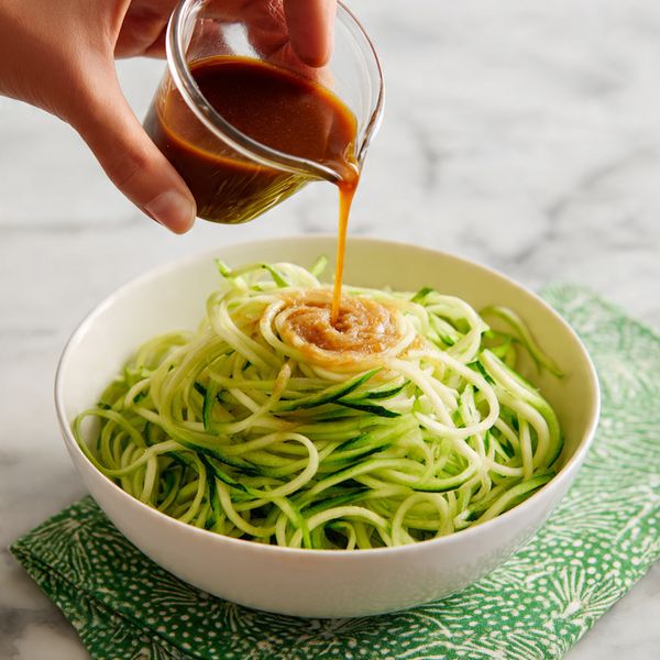 A white bowl is filled with a tall mound of light green spiralized cucumber noodles that look fresh and curly. On top, there is a small amount of a light brown paste or sauce in the center. Above the bowl, a woman's hand is pouring a thin stream of dark brown dressing from a small glass container onto the sauce and cucumber noodles. The bowl sits on a green patterned cloth, all set on a white marbled surface. photo taken with an iphone --ar 4:5 --v 7