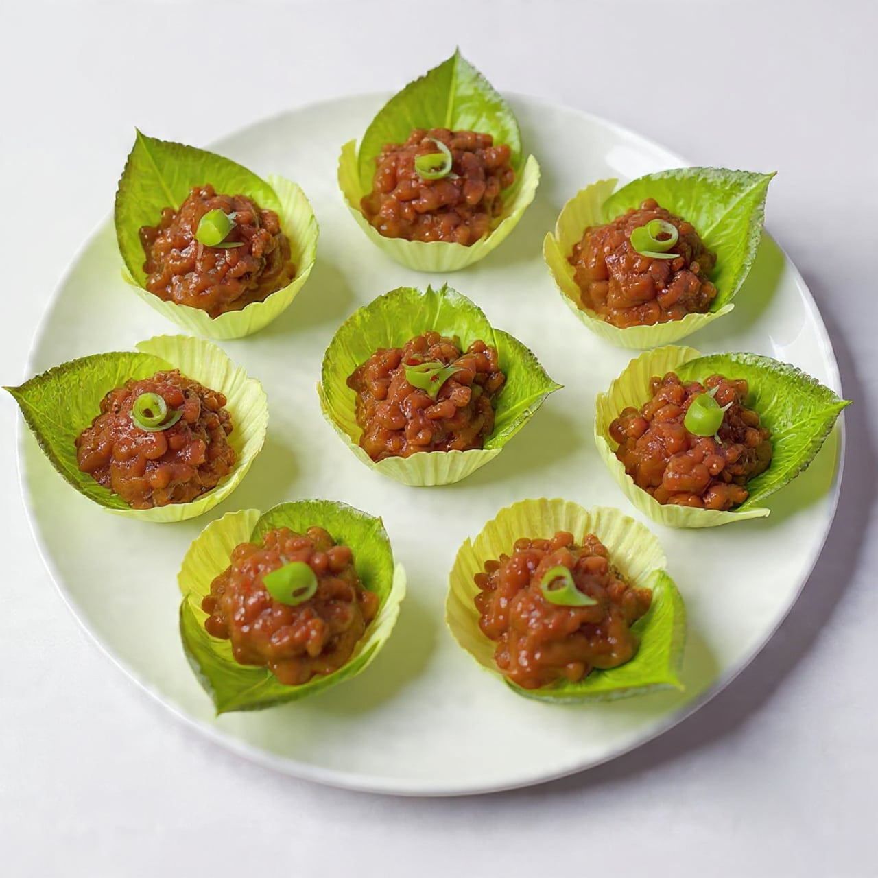 A white plate with five fresh green lettuce leaves arranged in a circle. Each leaf holds a dark brown, cooked mixture that looks like minced meat with small pieces and a slightly glossy texture. The mixture is sprinkled with small slices of green onions and bits of red chili, adding pops of green and red color on top. The plate sits on a white marbled surface. photo taken with an iphone --ar 4:5 --v 7