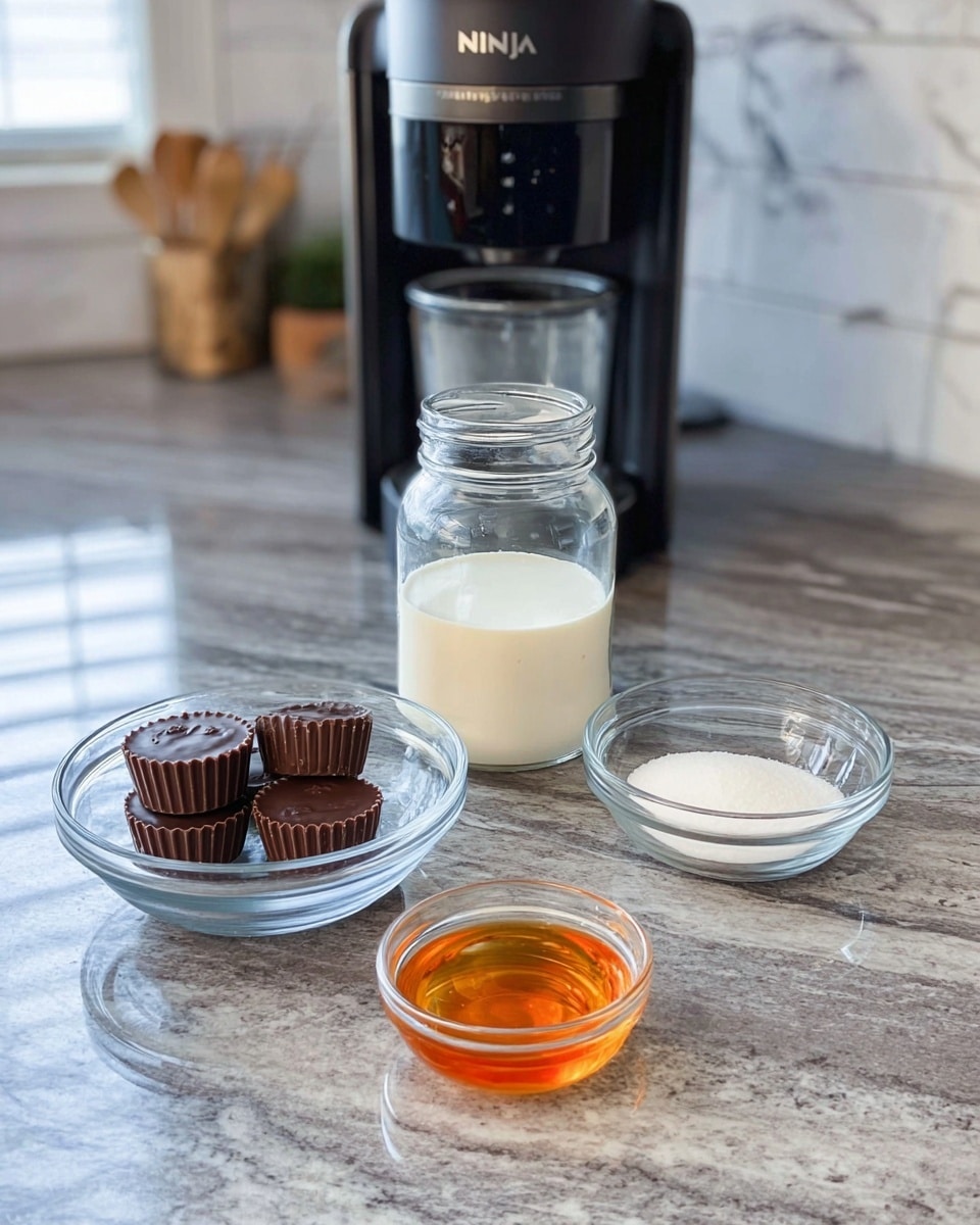 The image shows a kitchen countertop with a black Ninja appliance in the center background. In front of the appliance, there is a clear glass jar filled halfway with a light cream liquid. To the left of the jar, a clear glass bowl holds three chocolate peanut butter cups. To the right, another clear glass bowl contains white granulated sugar. In the front-center, a small white bowl contains a golden amber liquid. The countertop is a polished grey stone with a white marbled texture in the background, and the setting is bright and clean. Photo taken with an iphone --ar 4:5 --v 7