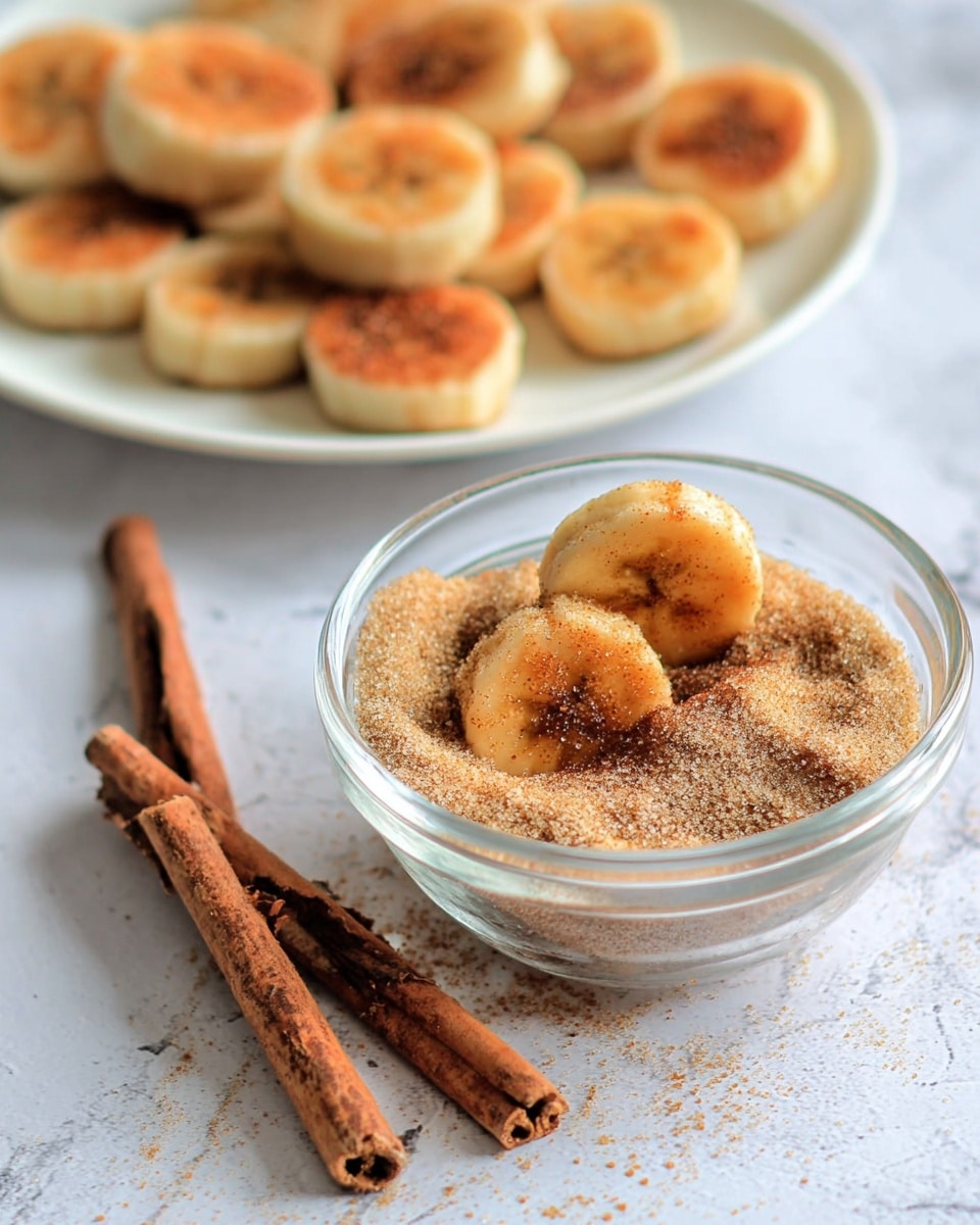 The image shows small round banana slices with a golden brown top on a white plate in the background, slightly blurred. In the front is a clear glass bowl filled with a cinnamon and sugar powder mix, where two banana slices are partially sunk in the powder and covered with it. Two cinnamon sticks lie next to the bowl on a soft white marbled surface. Another banana slice is placed on the surface close to the bowl. The overall colors include the warm brown of the cinnamon and bananas, the transparent bowl, and the white plate and surface. photo taken with an iphone --ar 4:5 --v 7