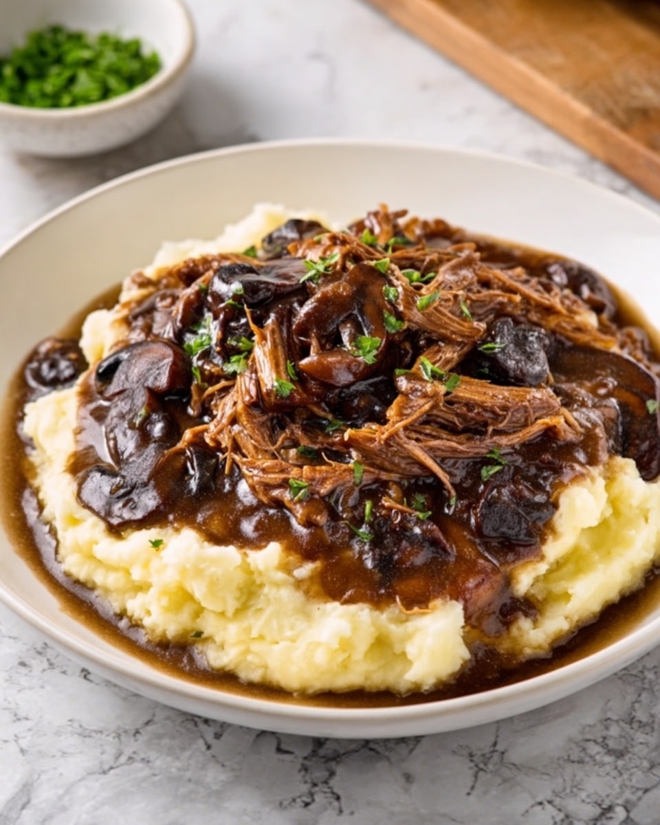 The image shows a white plate with a layer of soft, creamy mashed potatoes that are light yellow in color and have a smooth, slightly fluffy texture. On top, there is a thick, rich brown stew with shredded meat and chunks of dark mushrooms, giving a mix of deep brown and black tones with a glossy, thick sauce. The stew sits mostly in the middle of the mashed potatoes, slightly spreading out but not covering the edges. In the background, there is a small blurry bowl with chopped green herbs on a white marbled surface. photo taken with an iphone --ar 4:5 --v 7