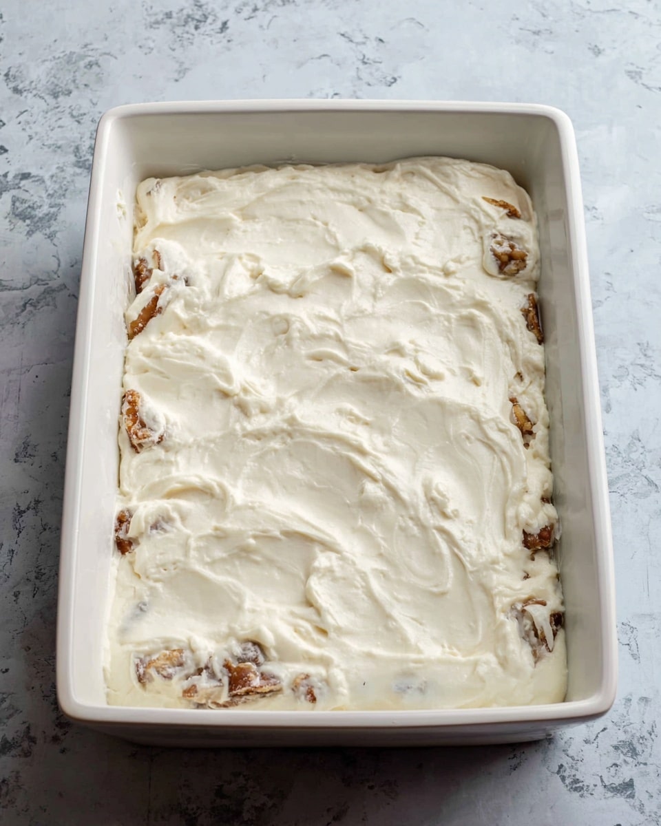 A white rectangular baking dish sits on a white marbled surface, filled with two visible layers; the bottom layer shows irregular chunks of light brown with some darker spots, likely cooked fruit or nuts, partially visible through the thick, creamy top layer. The top layer is smooth, white, and slightly textured with soft swirls and small lumps, covering the entire surface evenly. The edges of the dish are clean, and the background shows a muted grey and white pattern, making the dish the clear focus of the image. photo taken with an iphone --ar 4:5 --v 7