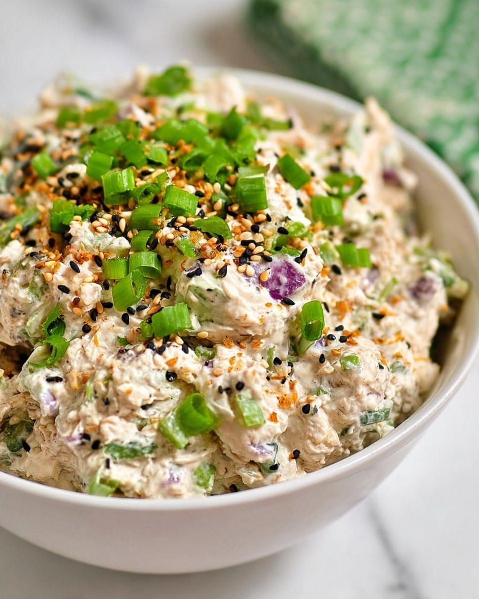 A white bowl filled with a creamy, chunky salad that has a light beige color base mixed with small pieces of purple, green, and white vegetables. On top, there are bright green chopped green onions scattered evenly, along with a sprinkle of black and tan seeds and small orange bits adding texture and color contrast. The bowl sits on a white marbled surface with a blurred green patterned object in the background. photo taken with an iphone --ar 4:5 --v 7