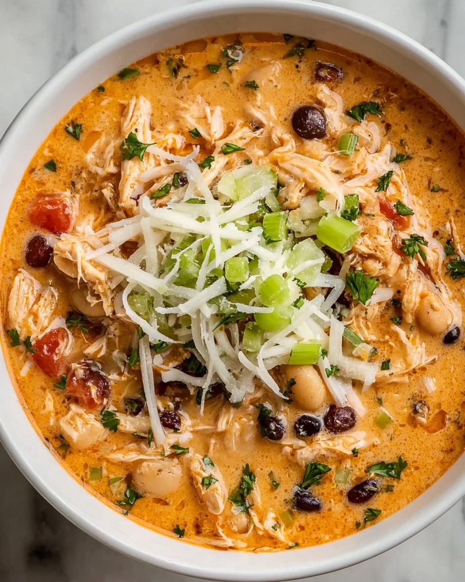 A close-up view of a creamy orange soup in a white bowl, filled with shredded chicken, white beans, black beans, and small tomato pieces, mixed in a rich, thick broth. On top of the soup, there is a layer of shredded white cheese and chopped green celery with smooth texture. Small bits of chopped white onion and green parsley are scattered across the surface, adding bursts of color to the creamy orange base. The bowl is placed on a white marbled texture surface. Photo taken with an iphone --ar 4:5 --v 7
