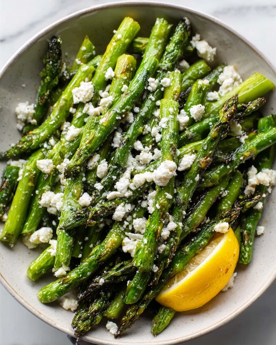 The image shows a white bowl filled with vibrant green asparagus spears that are cooked and slightly charred, placed in a random pile inside the bowl. On top, there is a sprinkling of soft white cheese crumbles scattered unevenly across the asparagus, adding a contrasting texture and color. To the side of the bowl, there is a bright yellow lemon wedge resting on the edge. The bowl is set on a white marbled surface, which gives a clean and light background to the dish. photo taken with an iphone --ar 4:5 --v 7