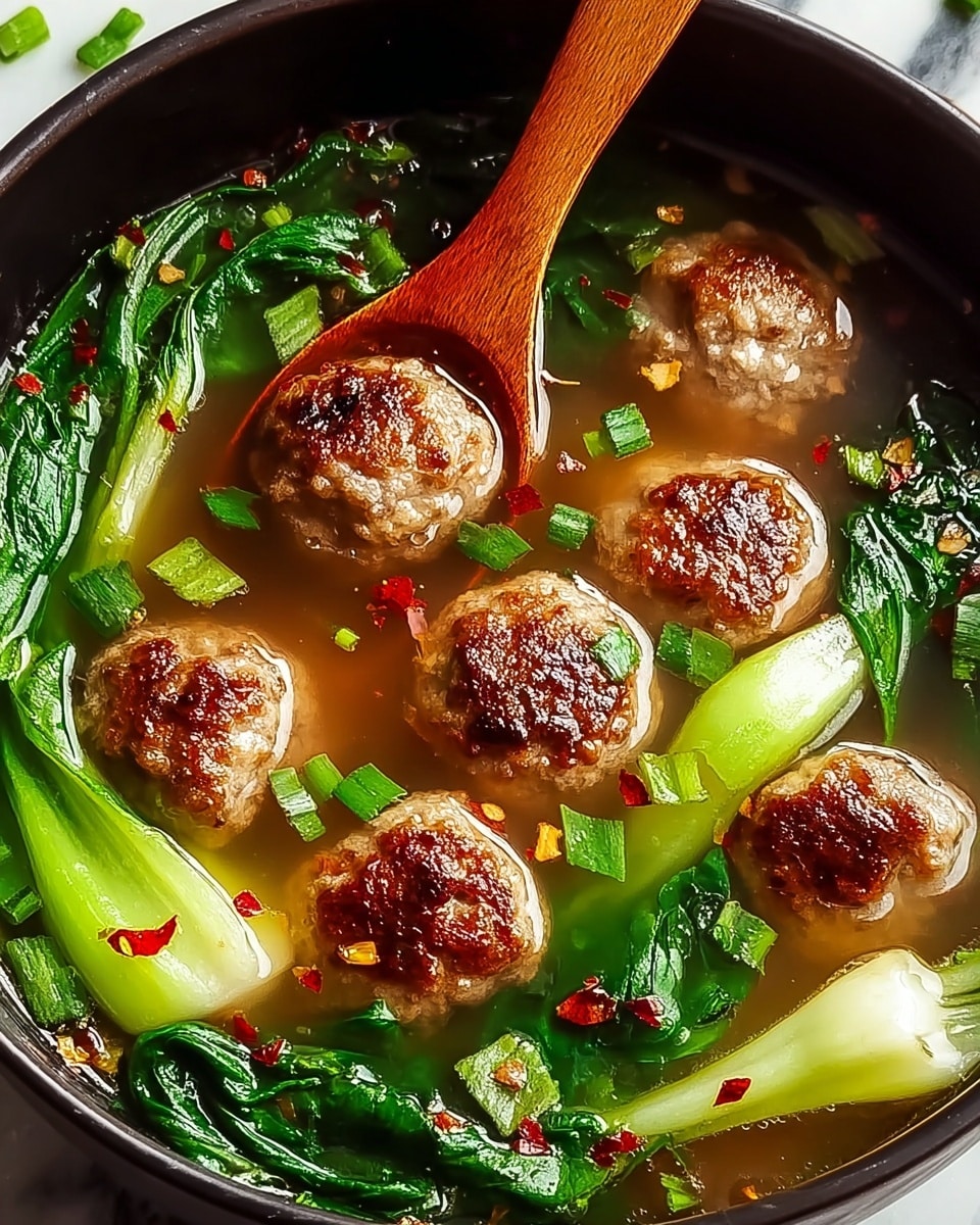 A close-up view of a dark bowl filled with clear broth soup containing six browned meatballs scattered throughout. Bright green bok choy leaves and stalks float around the meatballs, with small chopped green onions sprinkled on top. The soup surface is speckled with red chili flakes, adding color contrast. A wooden spoon rests inside the bowl, partially submerged in the broth, creating a natural warm tone. The entire bowl is placed on a white marbled surface. Photo taken with an iphone --ar 4:5 --v 7