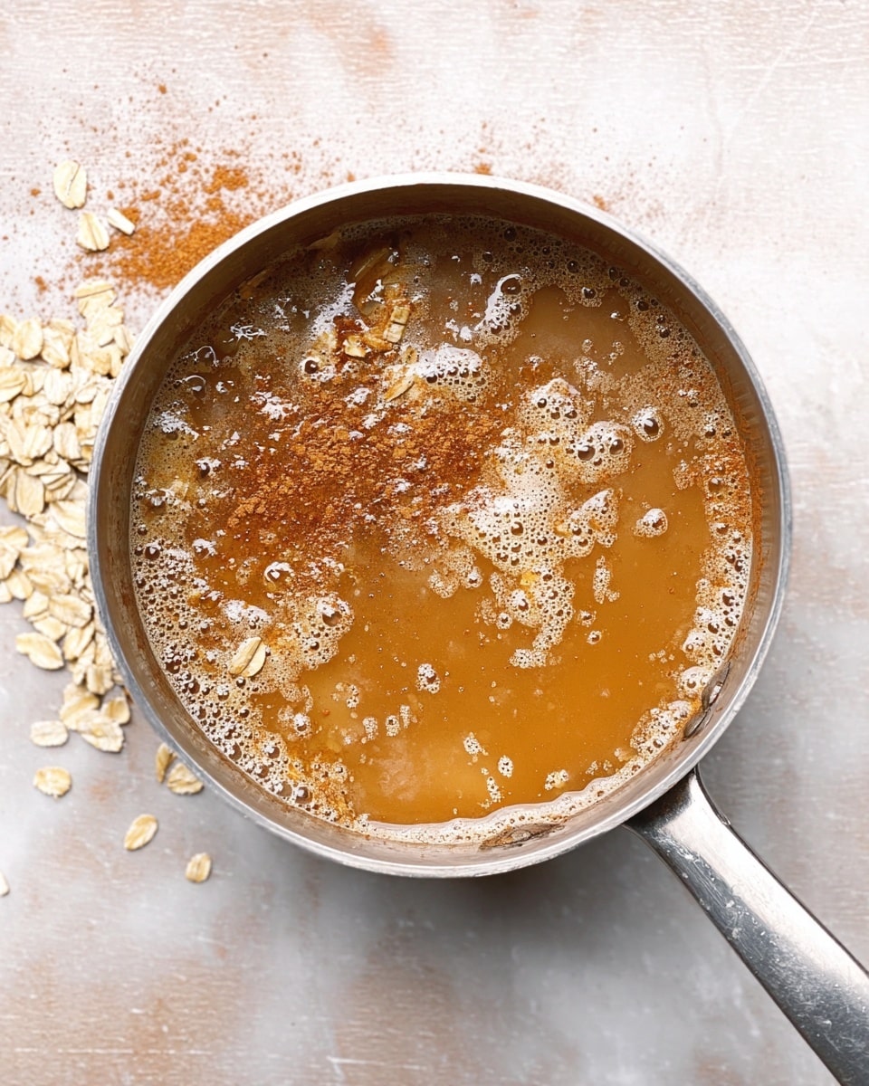 A top view of a silver pot filled with a light brown liquid that has cinnamon powder sprinkled on the surface, some foam bubbles, and scattered oat flakes. The pot is placed on a white marbled textured surface, and the metal handle of the pot is visible on the right side. photo taken with an iphone --ar 4:5 --v 7