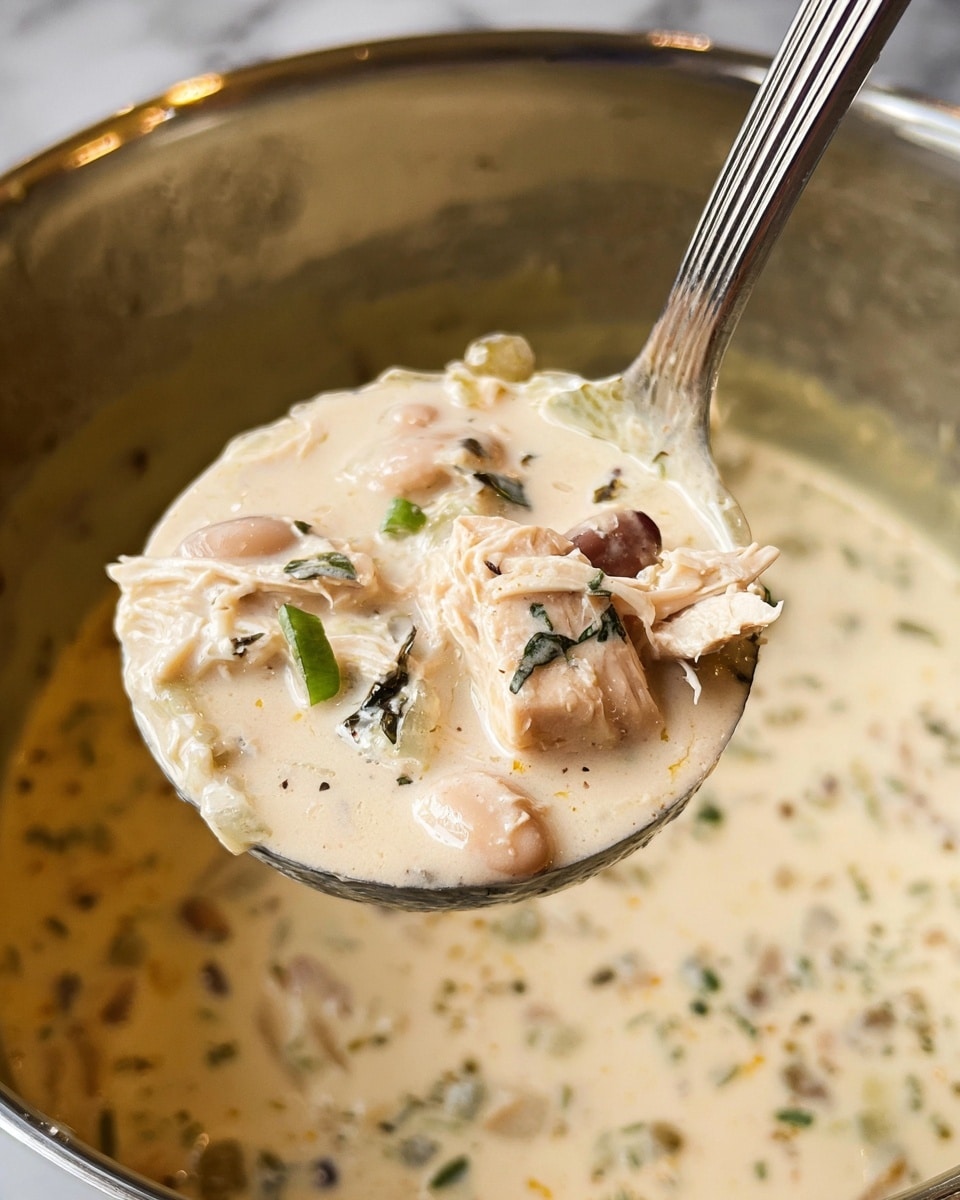 A close-up of a ladle filled with creamy soup showing three layers: a smooth, thick cream base with soft off-white color, mixed with chunks of light beige beans and small pieces of shredded white chicken, and sprinkled with dark green herbs. The soup has a slightly chunky texture with visible bits of finely chopped green onions and herbs scattered throughout. The ladle is shiny metal, reflecting parts of the white marbled background, and is held over a metal pot that contains more of the same creamy soup. The photo taken with an iphone --ar 4:5 --v 7