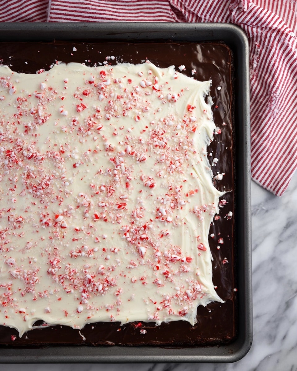The image shows a large rectangular tray filled with a layered dessert. The bottom layer is deep dark brown, smooth and glossy like chocolate. On top of that is a thick, uneven spread of white creamy layer sprinkled with small red and white crushed candy pieces scattered all over. The edges of the dessert are slightly rough, with some creamy white spread extending over the dark layer. The tray rests on a white marbled surface with a red and white striped cloth partially visible at the top. photo taken with an iphone --ar 4:5 --v 7