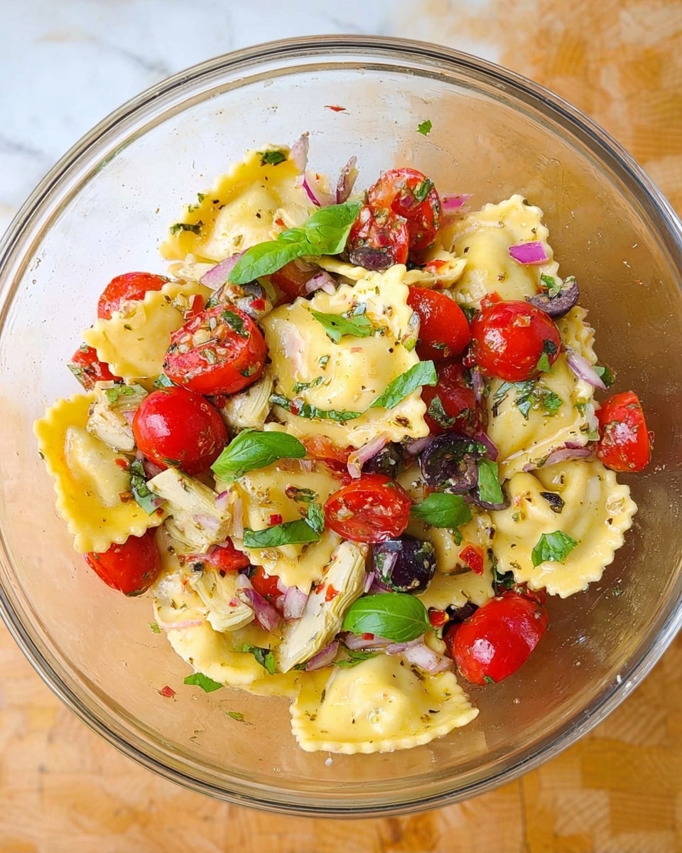 A clear glass bowl holds a fresh pasta salad layered with round, yellow ravioli pasta with ruffled edges, mixed with halved bright red cherry tomatoes, small green basil leaves scattered on top, pale artichoke hearts, chopped black olives, and bits of purple onion and red pepper. The ingredients are lightly coated with herbs and olive oil, all arranged on a wooden surface replaced by a white marbled texture. photo taken with an iphone --ar 4:5 --v 7