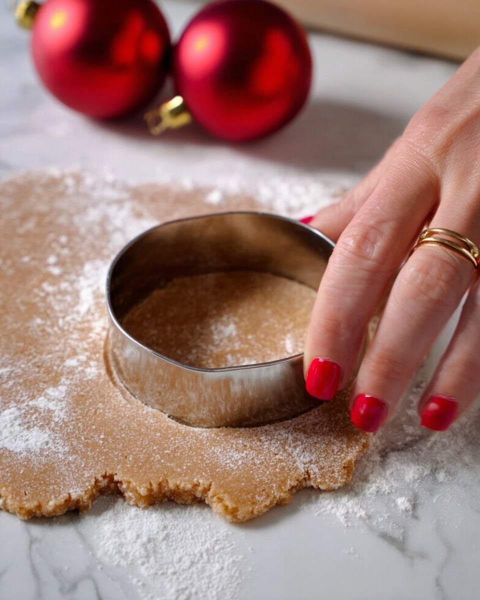A close-up view of a woman's hand with red nail polish and a gold ring pressing a round metal cookie cutter into light brown dough that has a dusting of white flour on top. The dough is laid flat on a white marbled surface. In the background, there are two shiny red Christmas ornaments adding a festive touch. Photo taken with an iphone --ar 4:5 --v 7
