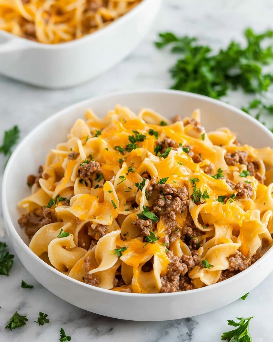 A white bowl filled with wide, curly egg noodles as the base layer, soft and pale yellow in color with a slight shine. Mixed throughout are small pieces of cooked ground beef in a light brown color, evenly spread over the noodles. On top of the noodles and beef is a melted layer of bright orange cheddar cheese, slightly stringy and blending into the noodles. Fresh green chopped parsley is sprinkled evenly over the dish, adding a fresh contrast. The bowl sits on a white marbled surface with some loose parsley leaves around it. In the background, a white dish with more of the same noodles and beef can be seen slightly out of focus. photo taken with an iphone --ar 4:5 --v 7