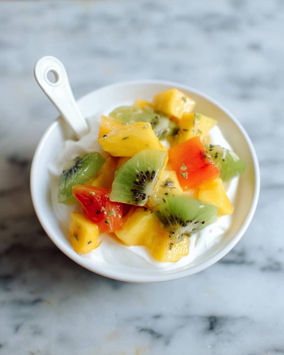 A white bowl filled with a layer of smooth white yogurt at the bottom, topped with chunks of colorful fruit including green kiwi with small black seeds, bright yellow mango pieces, and vibrant orange papaya. The fruits look fresh and juicy with a mix of soft and slightly firm textures. A white spoon with a looped handle stands in the bowl. The scene is set on a white marbled surface. photo taken with an iphone --ar 4:5 --v 7