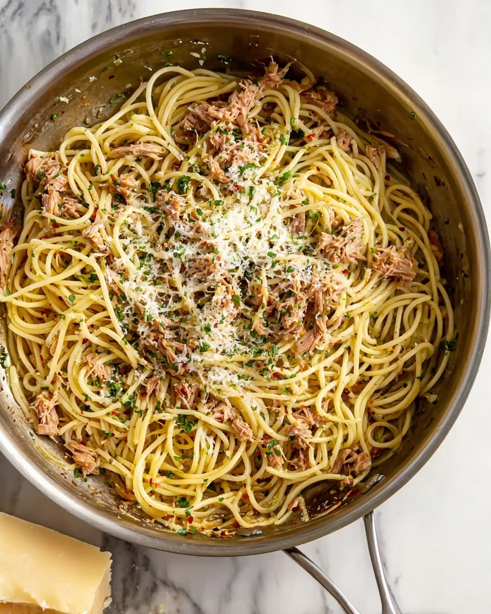 A stainless steel pan filled with a dish of spaghetti noodles layered throughout with small pieces of light brown tuna mixed evenly, sprinkled with finely chopped green herbs, and topped lightly with grated white cheese. The noodles are long and yellow, showing swirls and tangles throughout the pan, with small bits of red chili flakes scattered subtly on top. The pan handle extends toward the viewer, and the background is a white marbled surface with a partial look at a cheese wedge at the bottom left corner. Photo taken with an iphone --ar 4:5 --v 7