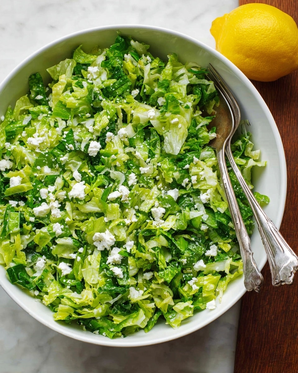 A white bowl filled with a fresh green salad showing two layers: the bottom layer is made of finely chopped mixed lettuce leaves in light and medium green shades with soft textures, and the top layer has small white cheese crumbles scattered evenly over the greens; a pair of silver forks rest inside the bowl on the right side, and a whole yellow lemon sits on a white marbled surface near the bowl’s bottom right corner, all under soft natural light. photo taken with an iphone --ar 4:5 --v 7