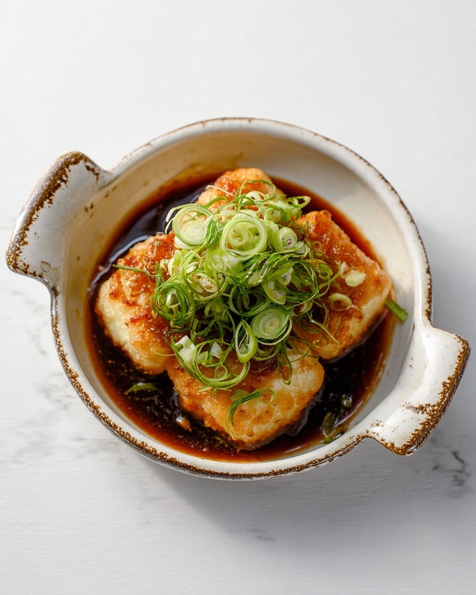 The dish has four pieces of light golden fried squares stacked in the center of a white bowl with handles on each side. The squares have a slightly crispy texture. Around and on top of the squares is a glossy dark brown sauce pooling in the bowl. Bright green thinly sliced scallions are sprinkled over the dish, adding a fresh look. The bowl is placed on a white marbled surface. Photo taken with an iphone --ar 4:5 --v 7