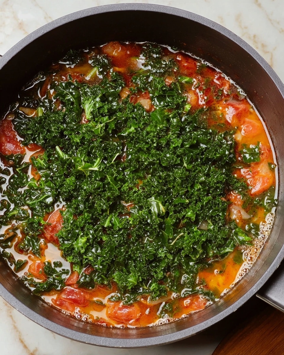 A close-up view of a large gray pot filled with a stew-like dish, layered with a bright orange soup base mixed with chunks of red tomatoes evenly spread around the edges, topped generously with a thick pile of dark green, curly chopped leafy greens covering the top center. The liquid looks rich and slightly oily, with small bubbles and a glossy texture visible on the surface. The pot is placed on a white marbled surface, adding contrast to the colors inside the pot. photo taken with an iphone --ar 4:5 --v 7