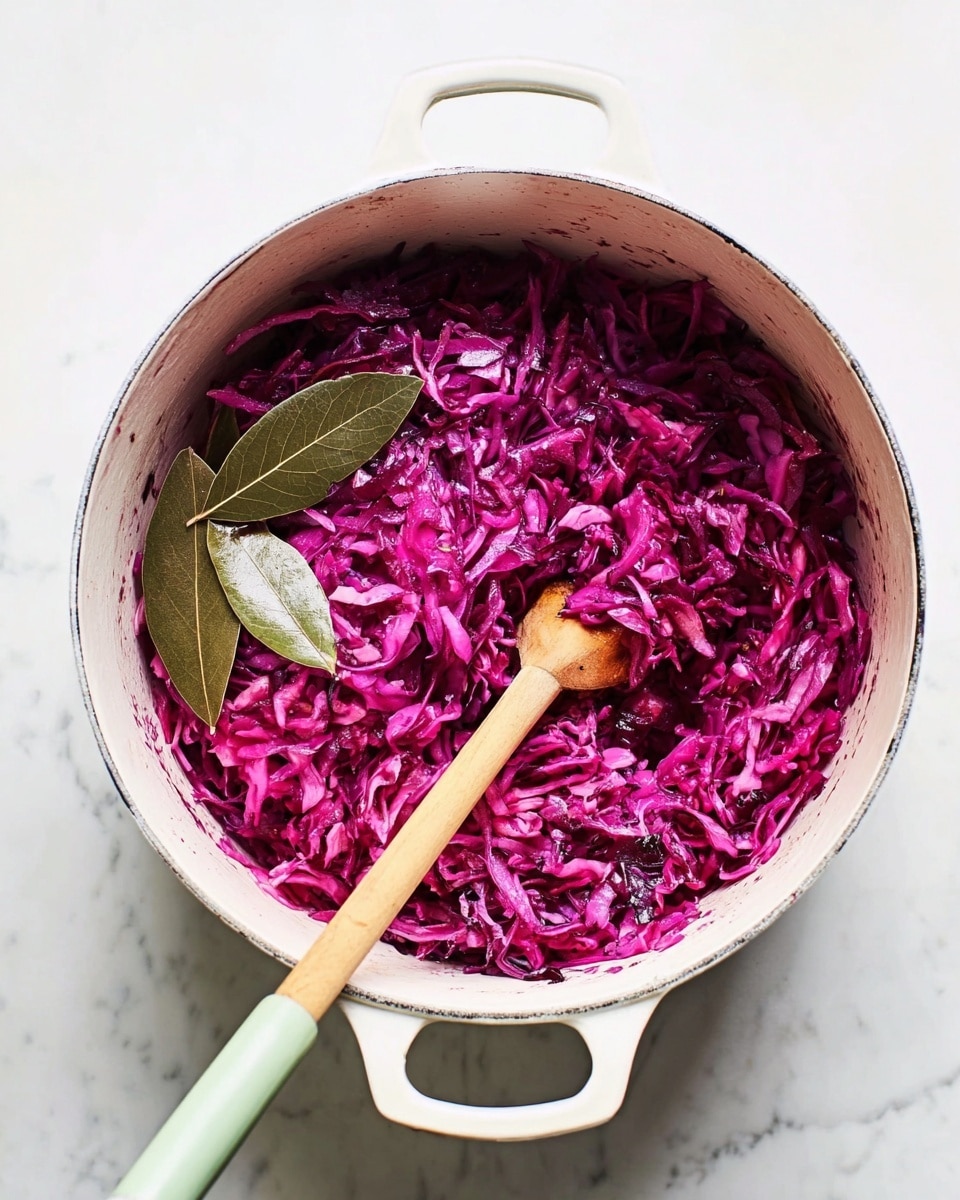 A white pot filled with cooked shredded red cabbage, showing its deep purple and pink colors with a slightly soft texture. Mixed in are two whole bay leaves on the left side of the pot. A wooden spoon with a pale green spatula end rests inside the pot, stirring the cabbage. The pot sits on a white marbled surface with clean, soft light highlighting the rich colors of the cabbage. photo taken with an iphone --ar 4:5 --v 7