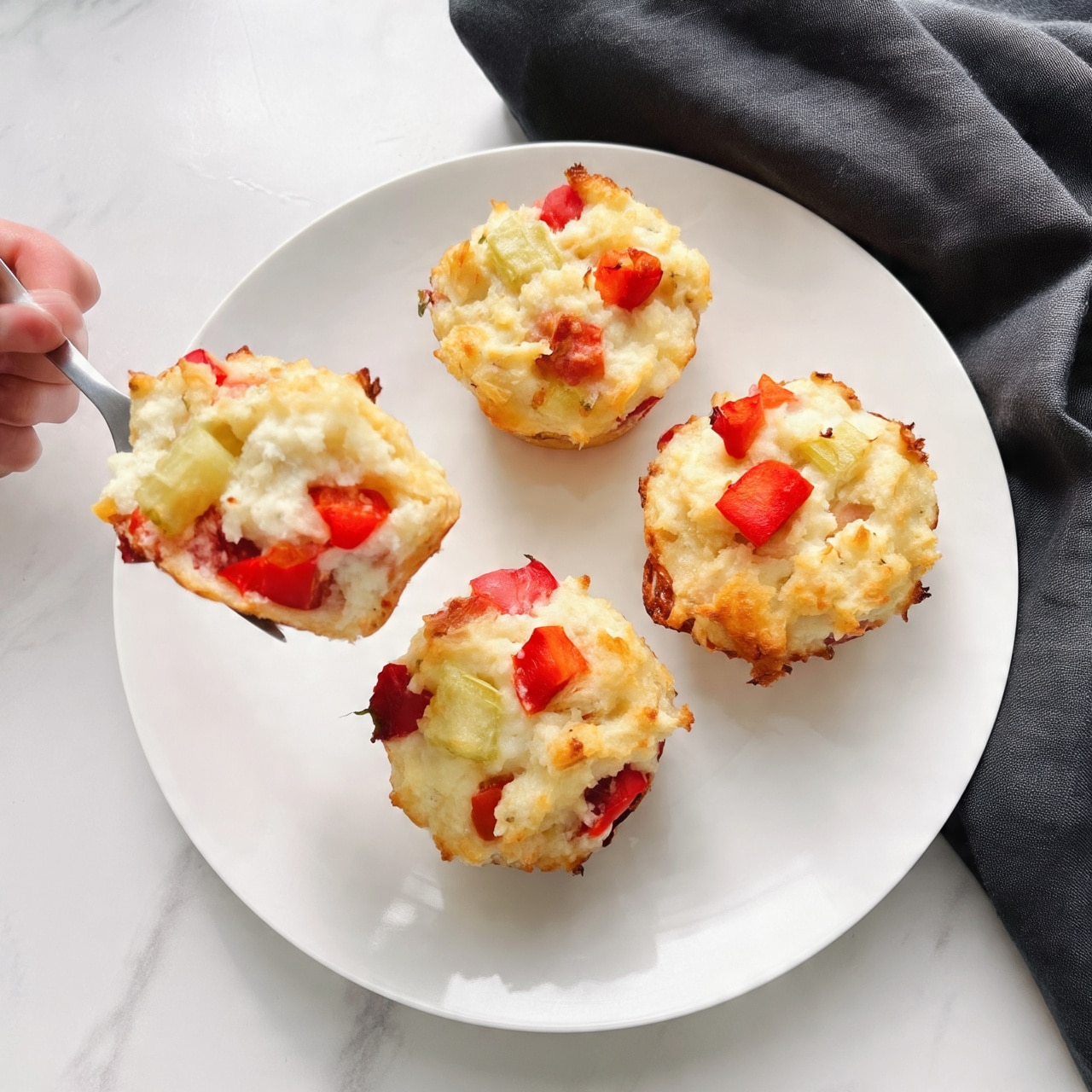 The image shows four small savory muffins on a white plate placed on a white marbled surface. Each muffin has a rough, slightly crumbly texture with visible chunks of red and green vegetables. The muffins have a pale golden color with bits of bright red peppers and light green celery scattered on top and throughout. A woman's hand is holding a fork that lifts one muffin, highlighting its soft, moist inside filled with the same colorful vegetable pieces. There is a dark gray cloth napkin next to the plate. photo taken with an iphone --ar 4:5 --v 7