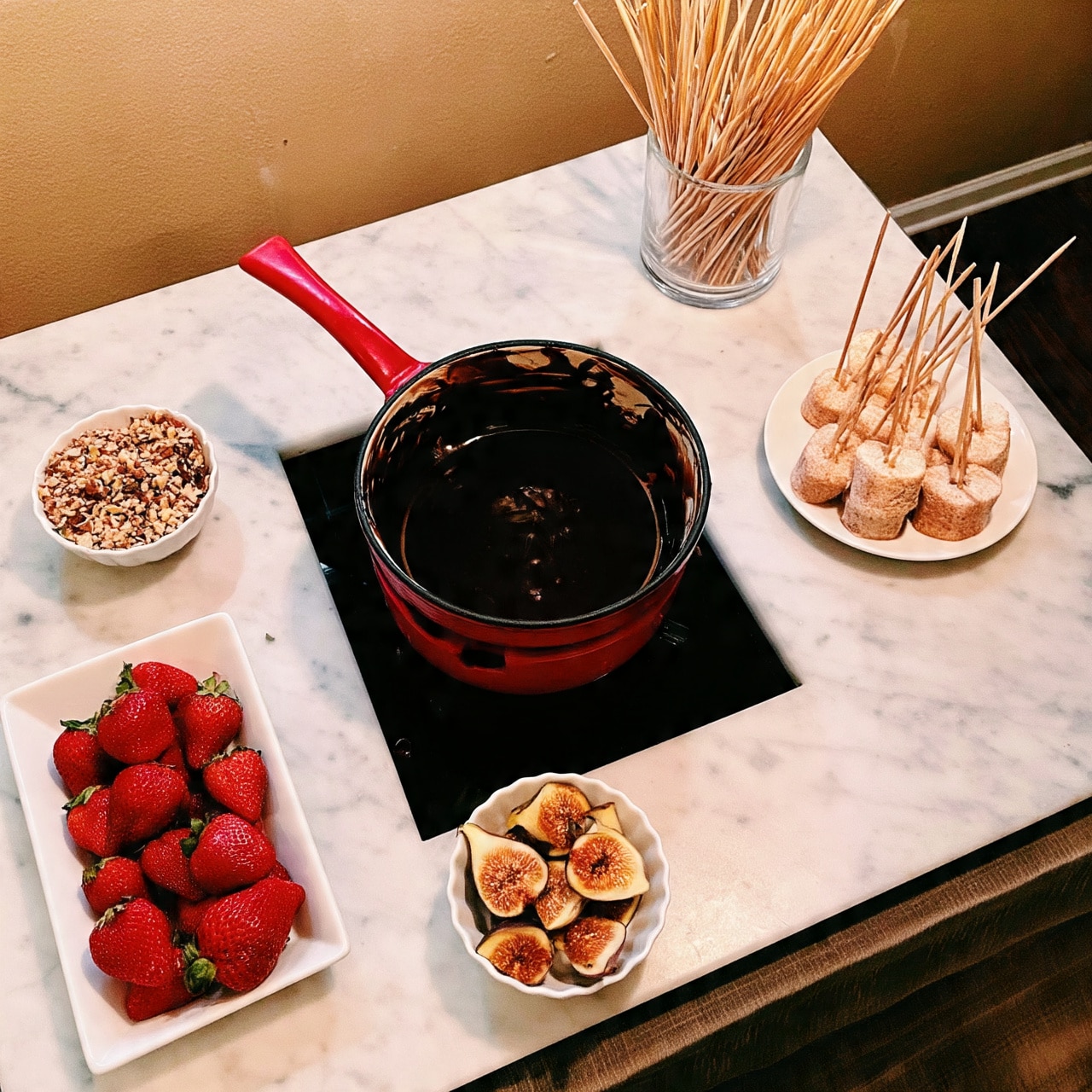 The image shows a red fondue pot filled with dark melted chocolate sitting on a white marble surface, with a black base underneath. Around the pot, there are several white dishes: one rectangular plate with bright red strawberries, a small bowl with chopped nuts, a round plate holding light-colored marshmallows on skewers, and a small dish containing halved figs. There is also a clear container holding wooden skewers upright in the background. The scene looks cozy and inviting, perfect for a chocolate fondue party. photo taken with an iphone --ar 4:5 --v 7