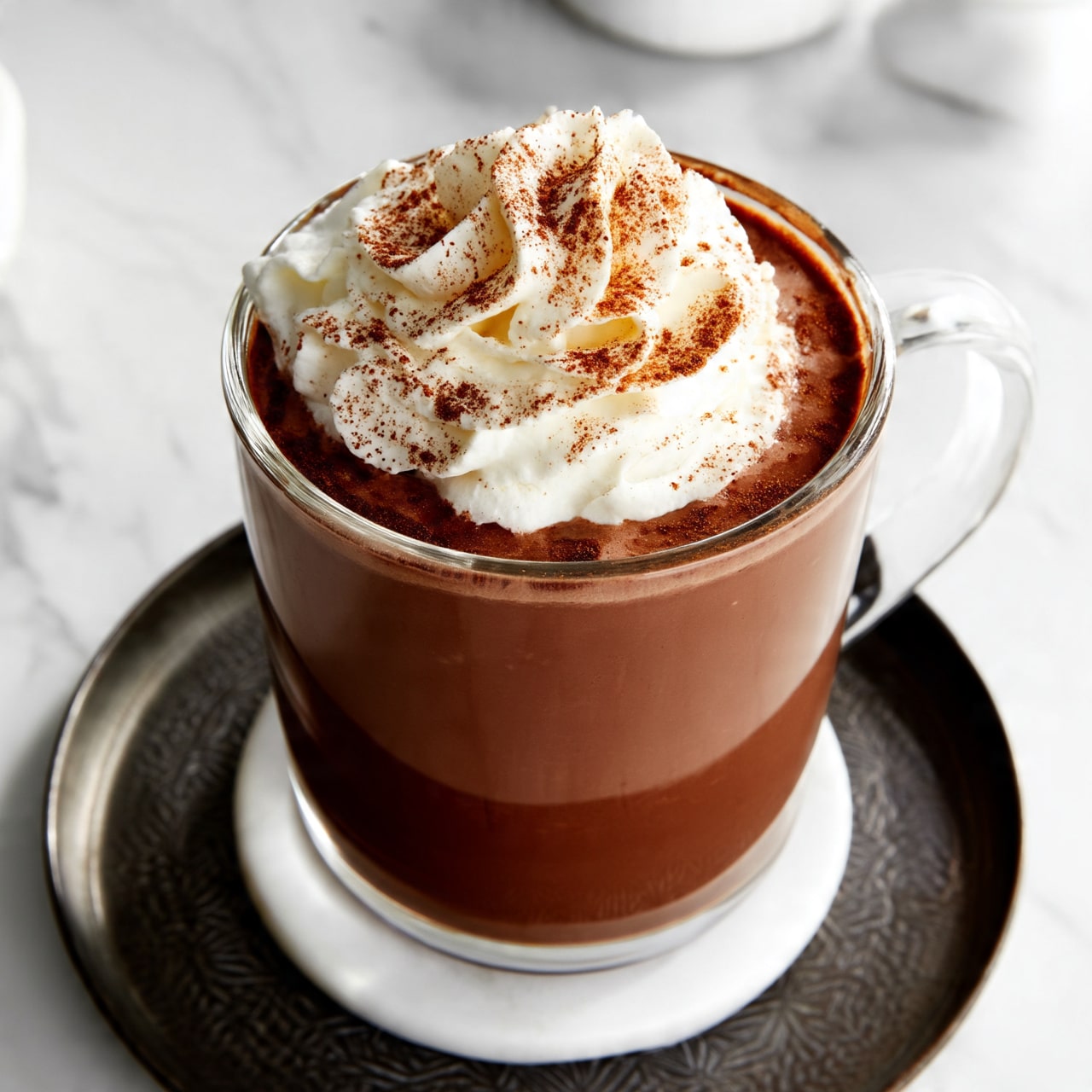 A clear glass mug filled with a thick brown hot chocolate drink, topped with a large swirl of white whipped cream dusted with fine brown cinnamon powder; the mug sits on a white coaster, which is placed on a round dark metal tray with a simple textured pattern, all set on a white marbled surface with soft lighting; in the background, blurred white elements add softness to the scene photo taken with an iphone --ar 4:5 --v 7